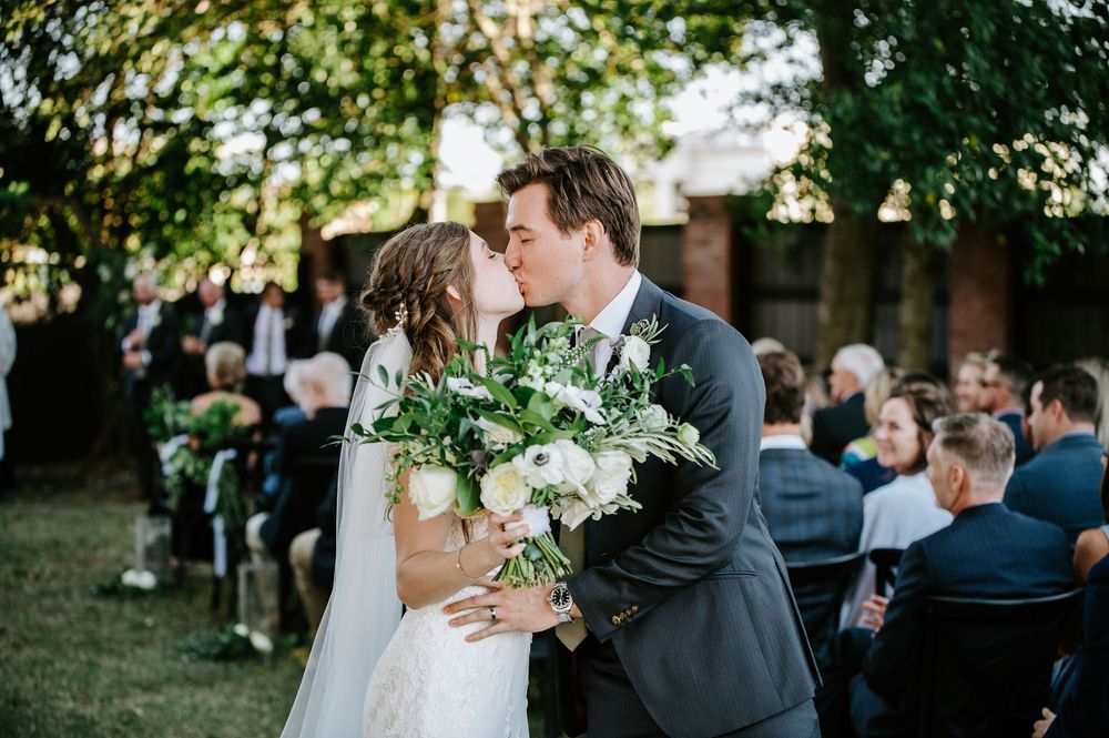 A couple shares a romantic kiss during an outdoor garden wedding ceremony surrounded by guests and greenery.