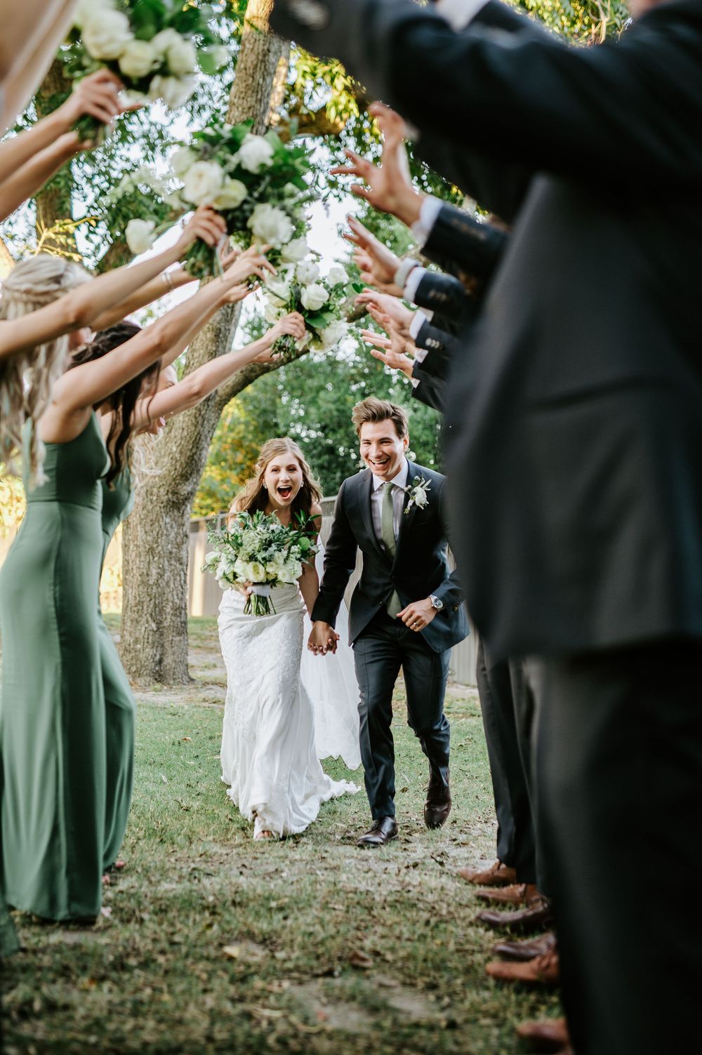 Wedding party creates tunnel formation outdoors with bridesmaids in sage green dresses.