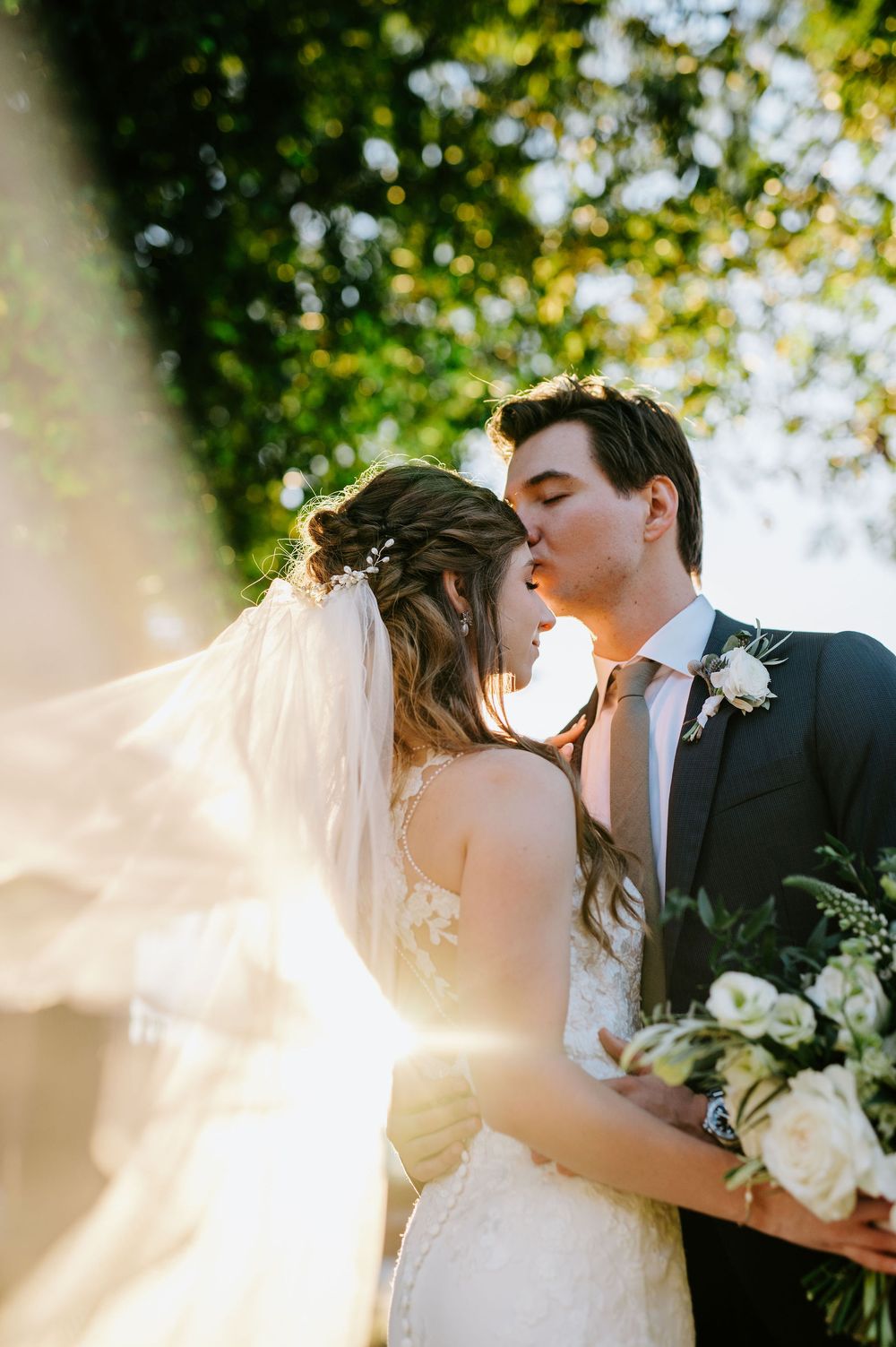 Romantic wedding couple sharing an intimate kiss in golden sunlight with flowing veil and white bouquet.