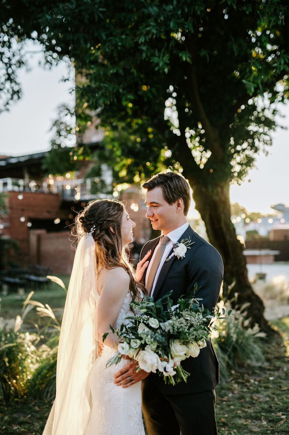 A romantic wedding couple embraces under a tree during golden hour sunset in an outdoor garden setting.