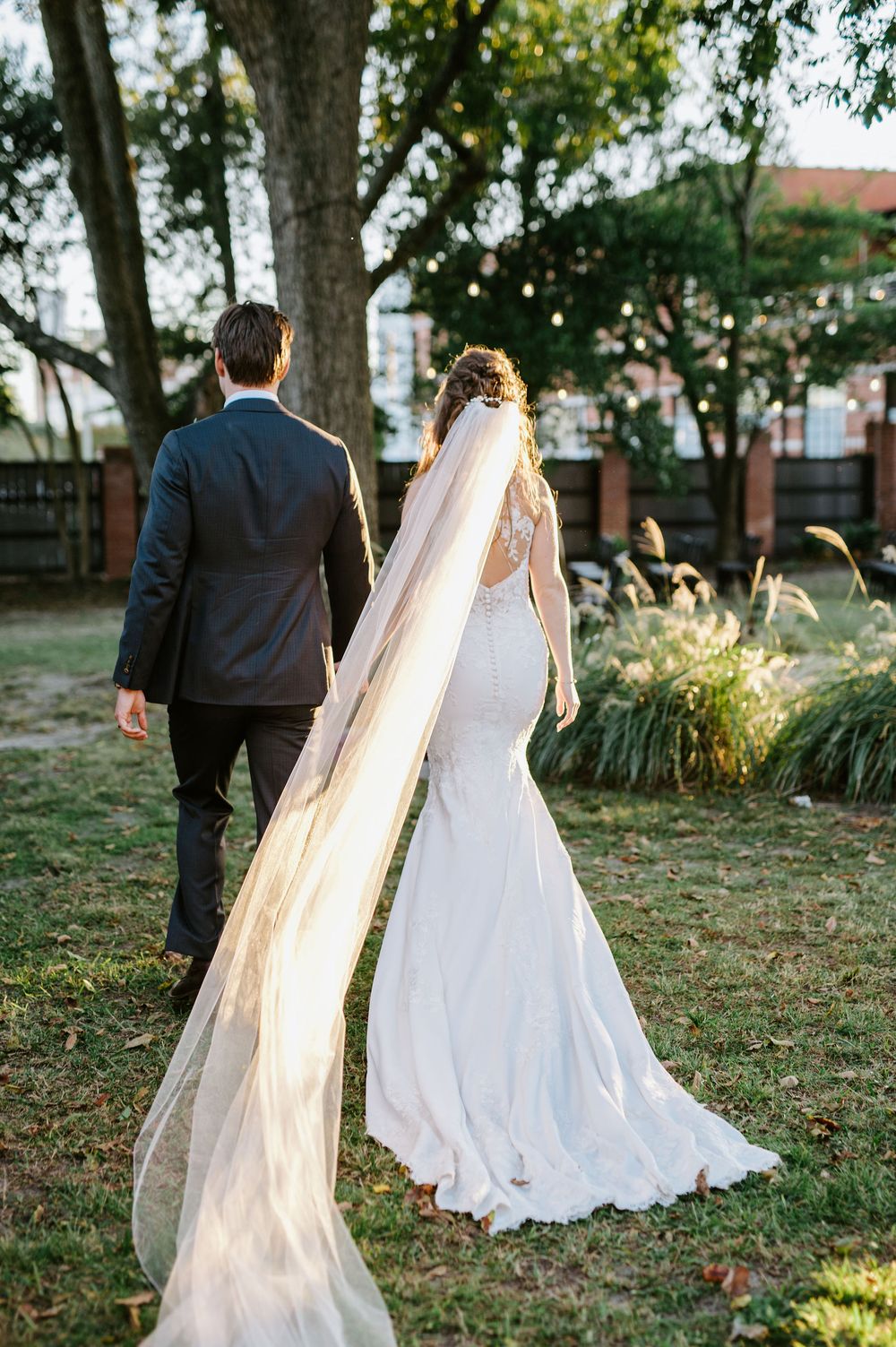Bride and groom walk hand in hand through a garden setting with sunlight filtering through trees.