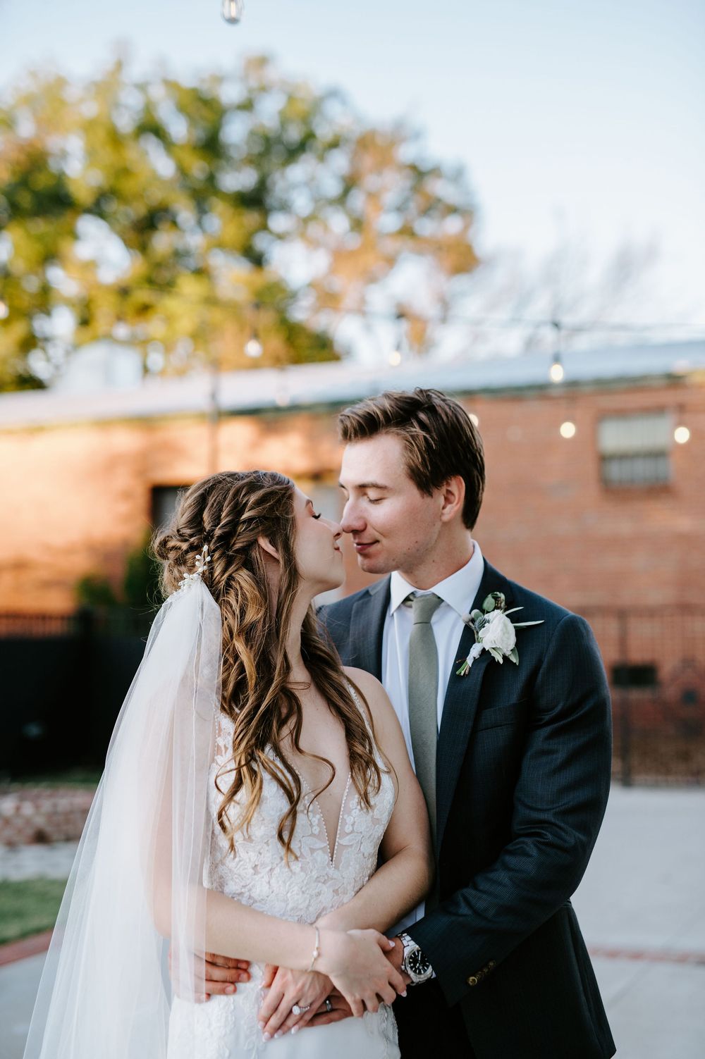 Newlyweds embrace in an outdoor wedding portrait against a brick wall with beautiful natural lighting.