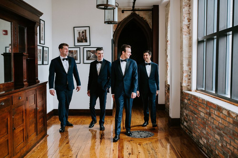 Groomsmen in dark suits gather in an elegant hallway with wooden details and natural light.