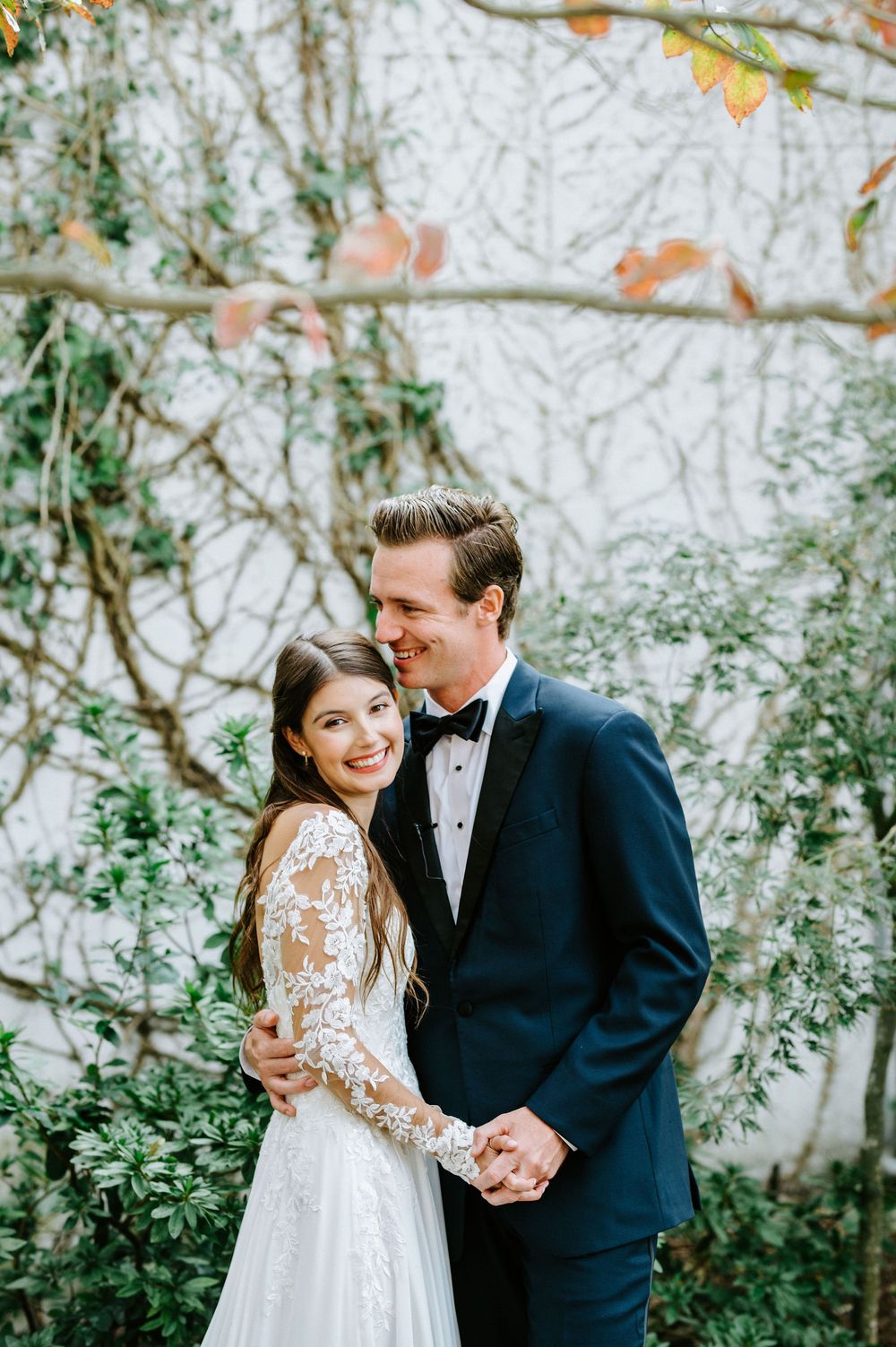 A romantic wedding portrait of a couple embracing outdoors with autumn foliage and greenery in the background.