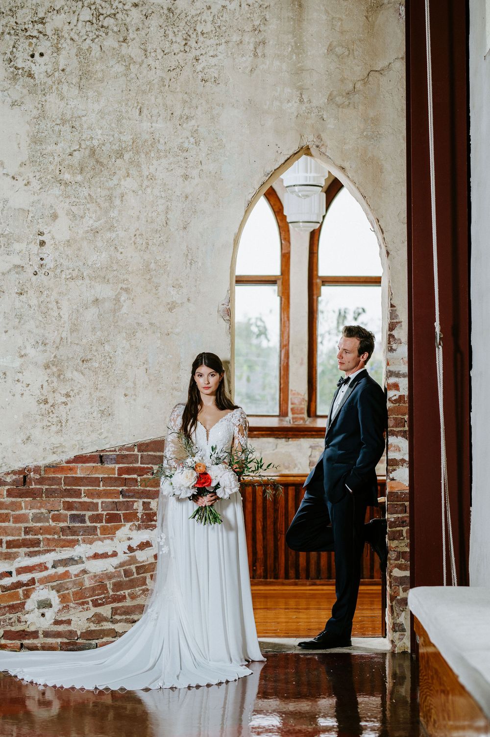 A couple poses in a rustic chapel with exposed brick walls and an arched gothic window creating dramatic lighting.
