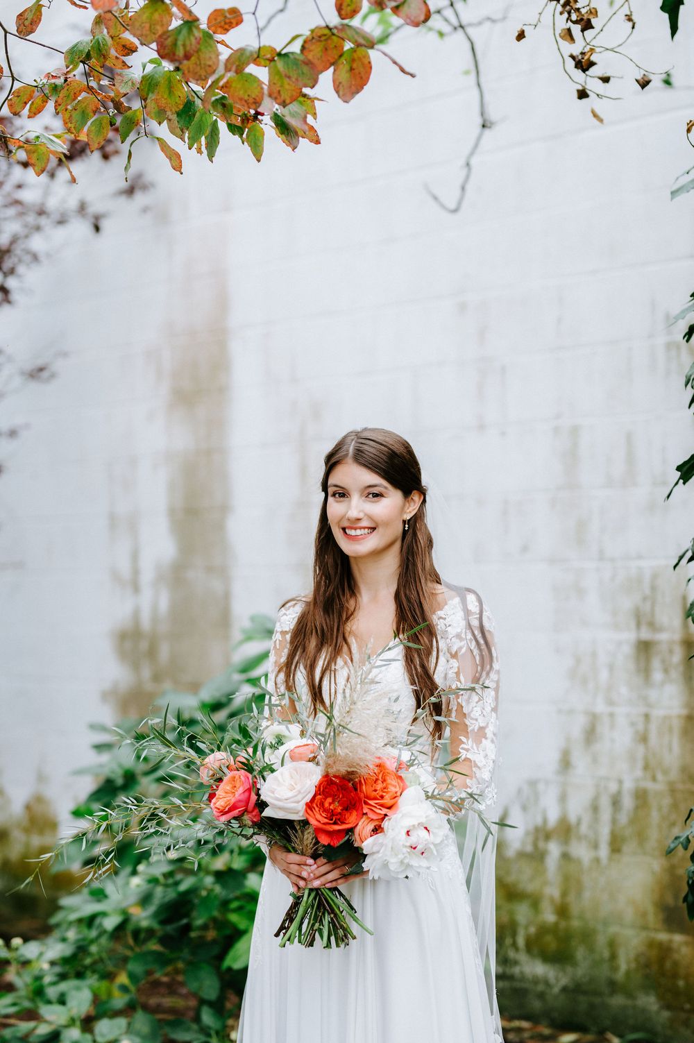 Bride in long-sleeved lace wedding dress holds vibrant coral and red rose bouquet against white stone wall with autumn leaves.