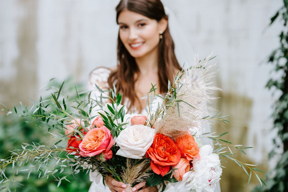 A vibrant bridal bouquet features coral roses, white blooms and wispy greenery in a romantic outdoor setting.