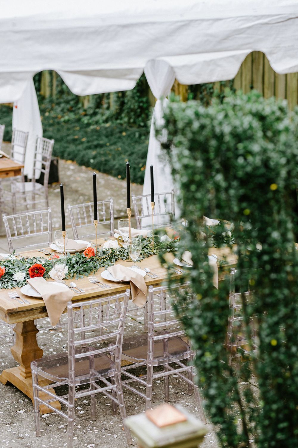 Elegant outdoor wedding reception setup with rustic wooden tables and clear chairs under white fabric draping.