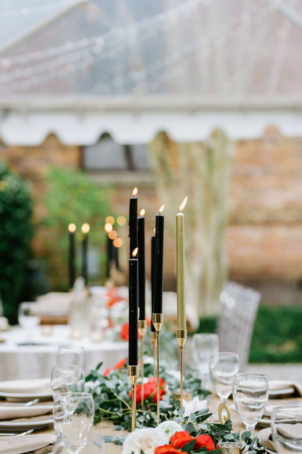 Elegant table setting with tall black and gold candles, greenery, and coral flowers under a white tent at outdoor event.
