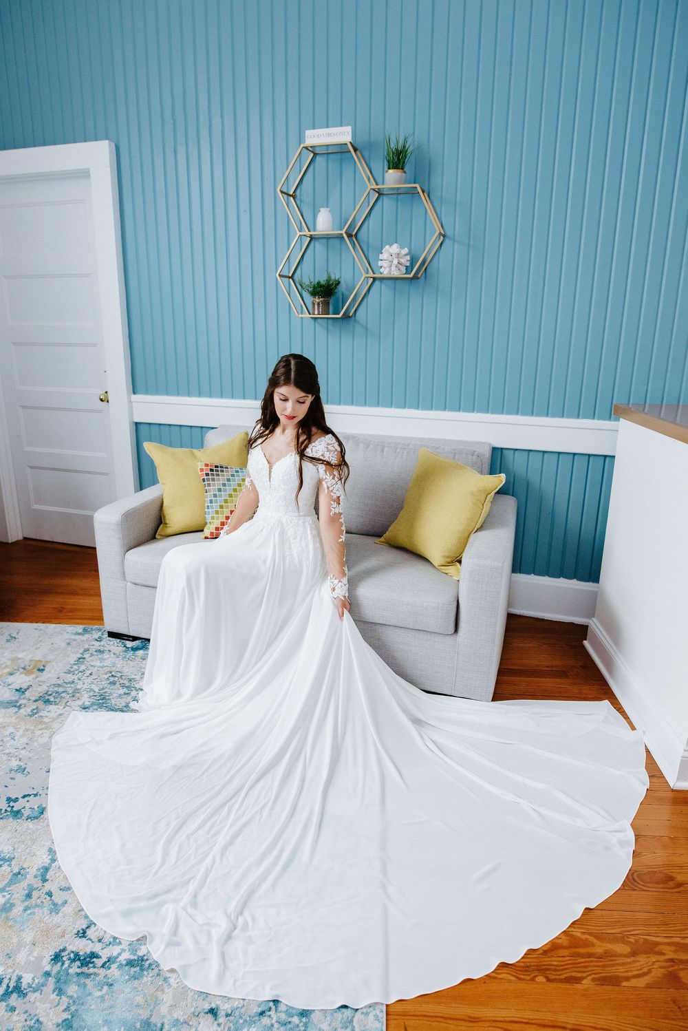 Elegant white wedding dress with flowing train displayed in a room with blue walls and hexagonal wall decor.