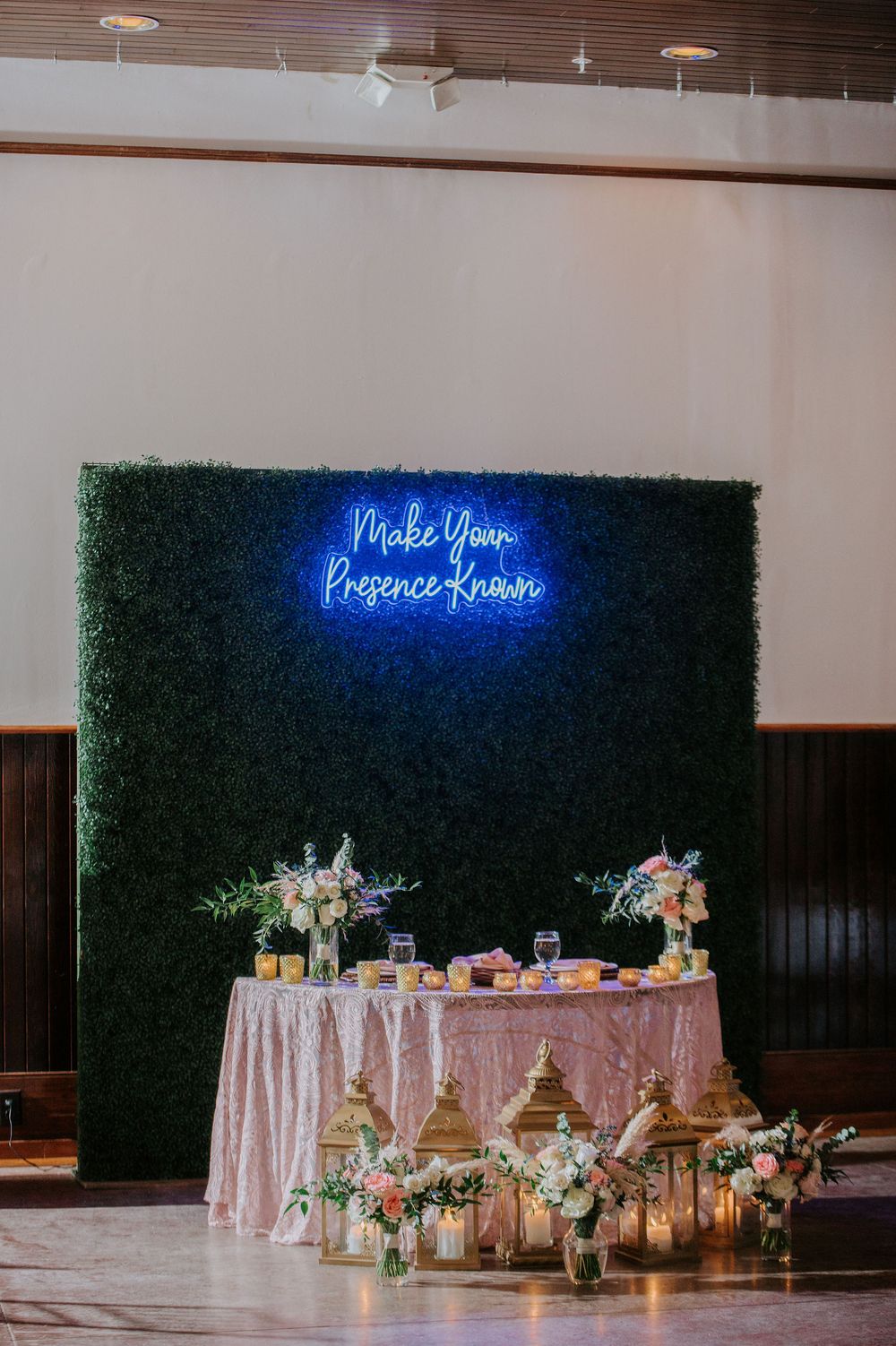 A wedding dessert table setup with pink tablecloth and greenery wall backdrop illuminated by blue neon signage.