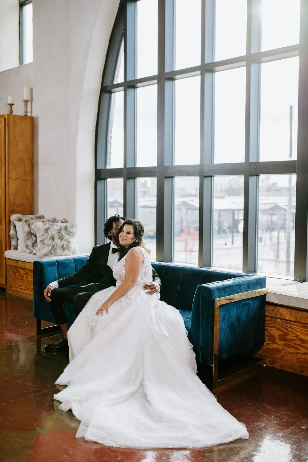 A bride in a flowing white wedding dress lounges on a blue velvet couch near large windows in an elegant room.