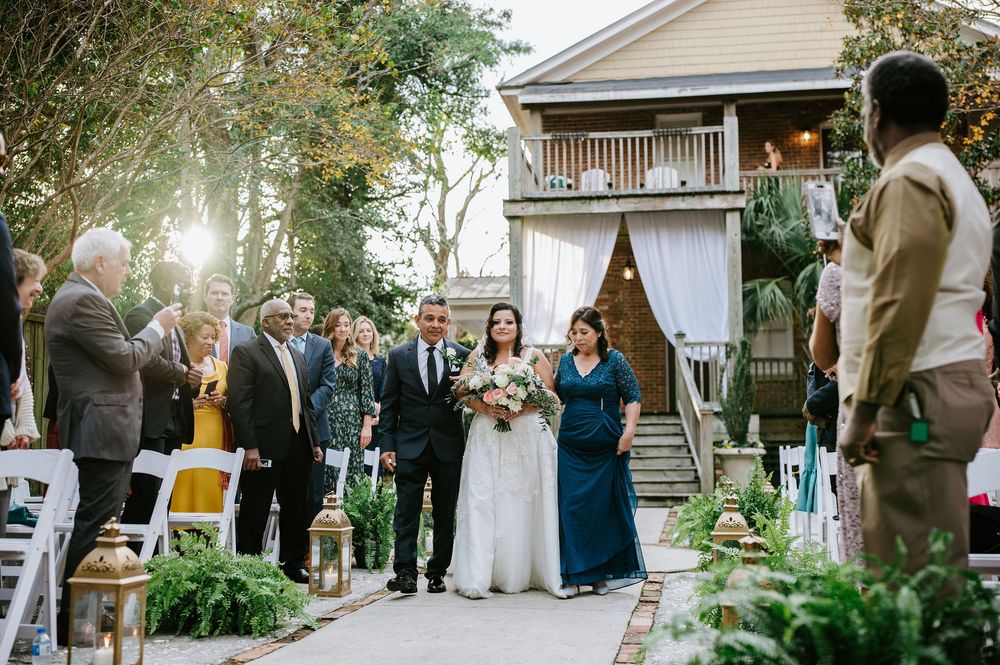 Guests gather for an outdoor wedding ceremony at a rustic venue with white wooden columns and lush greenery.