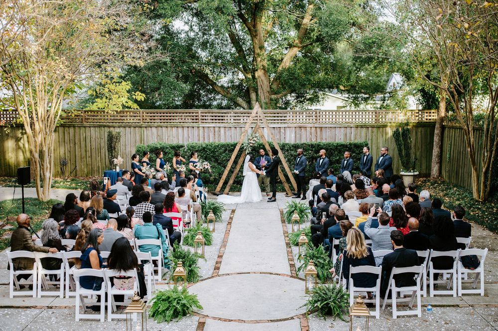 Outdoor garden wedding ceremony with white chairs and greenery lining the aisle under a wooden triangle arch.