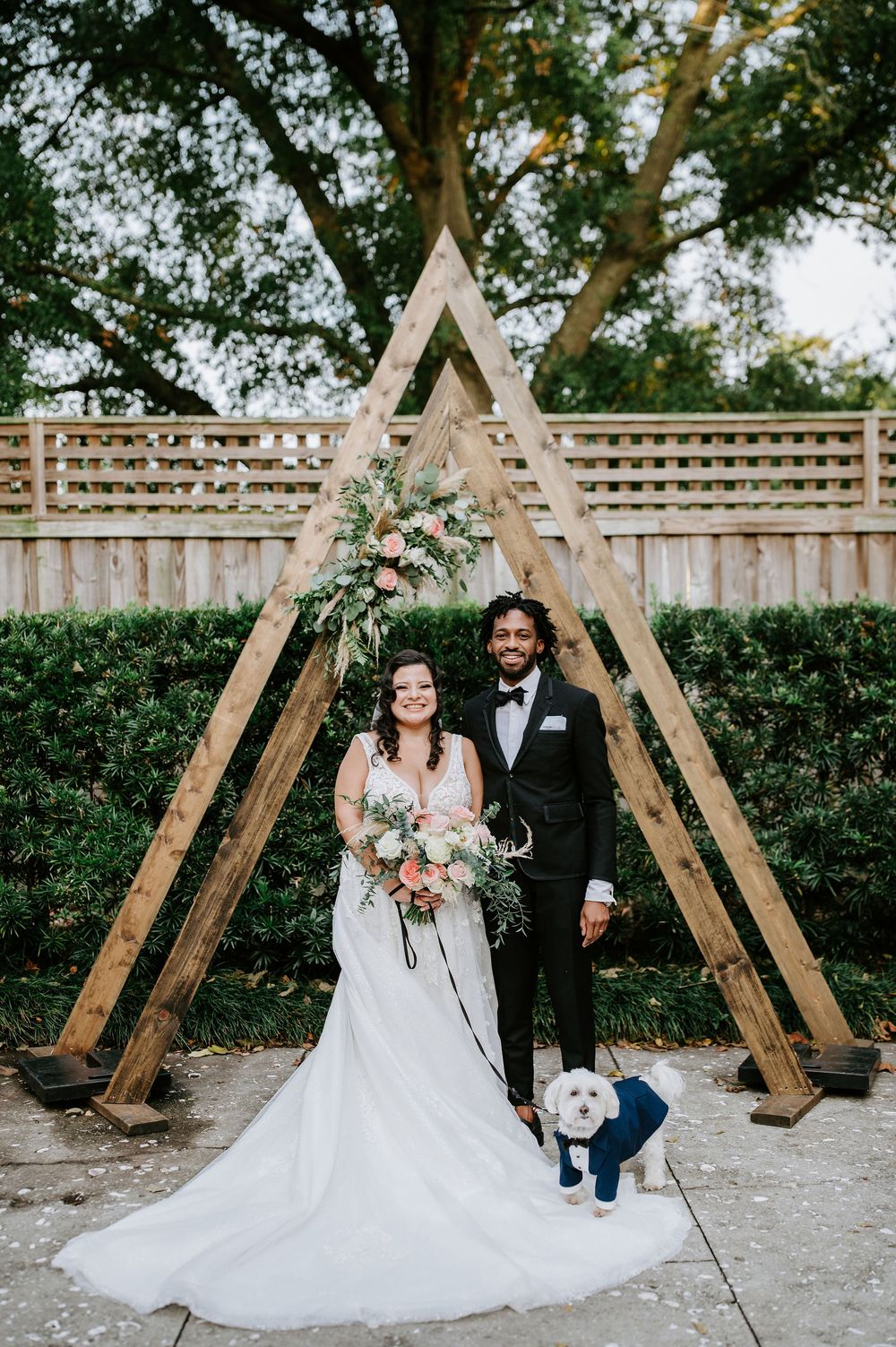 Wooden A-frame wedding arch decorated with floral arrangements stands against a garden hedge backdrop for an outdoor ceremony.