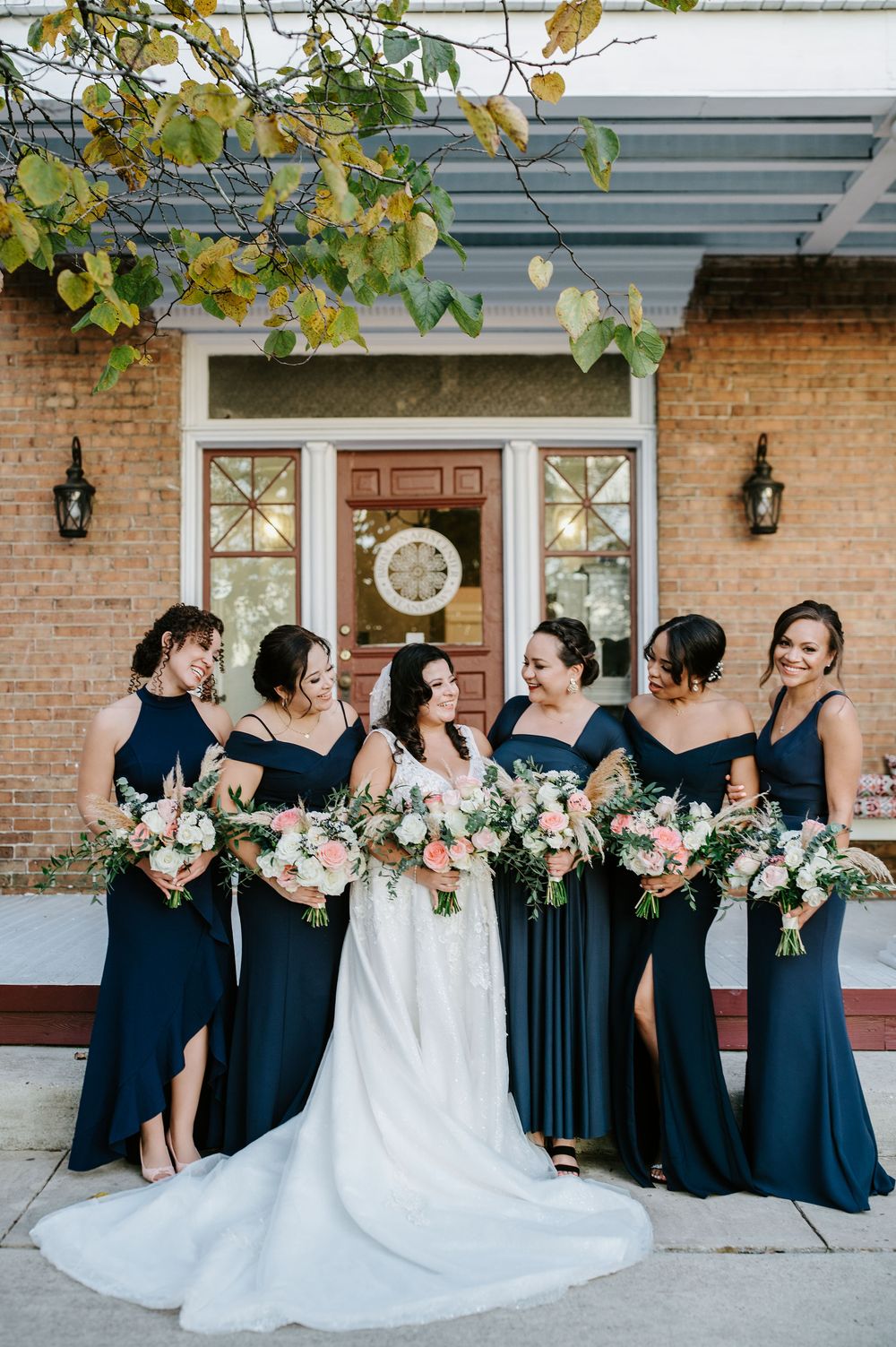 A bridal party in navy blue dresses and white bouquets poses together on a brick porch.