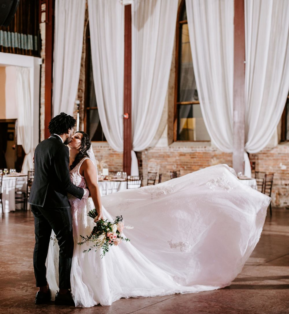 Bride and groom share an intimate moment in a rustic venue with flowing white curtains and dramatic lighting.