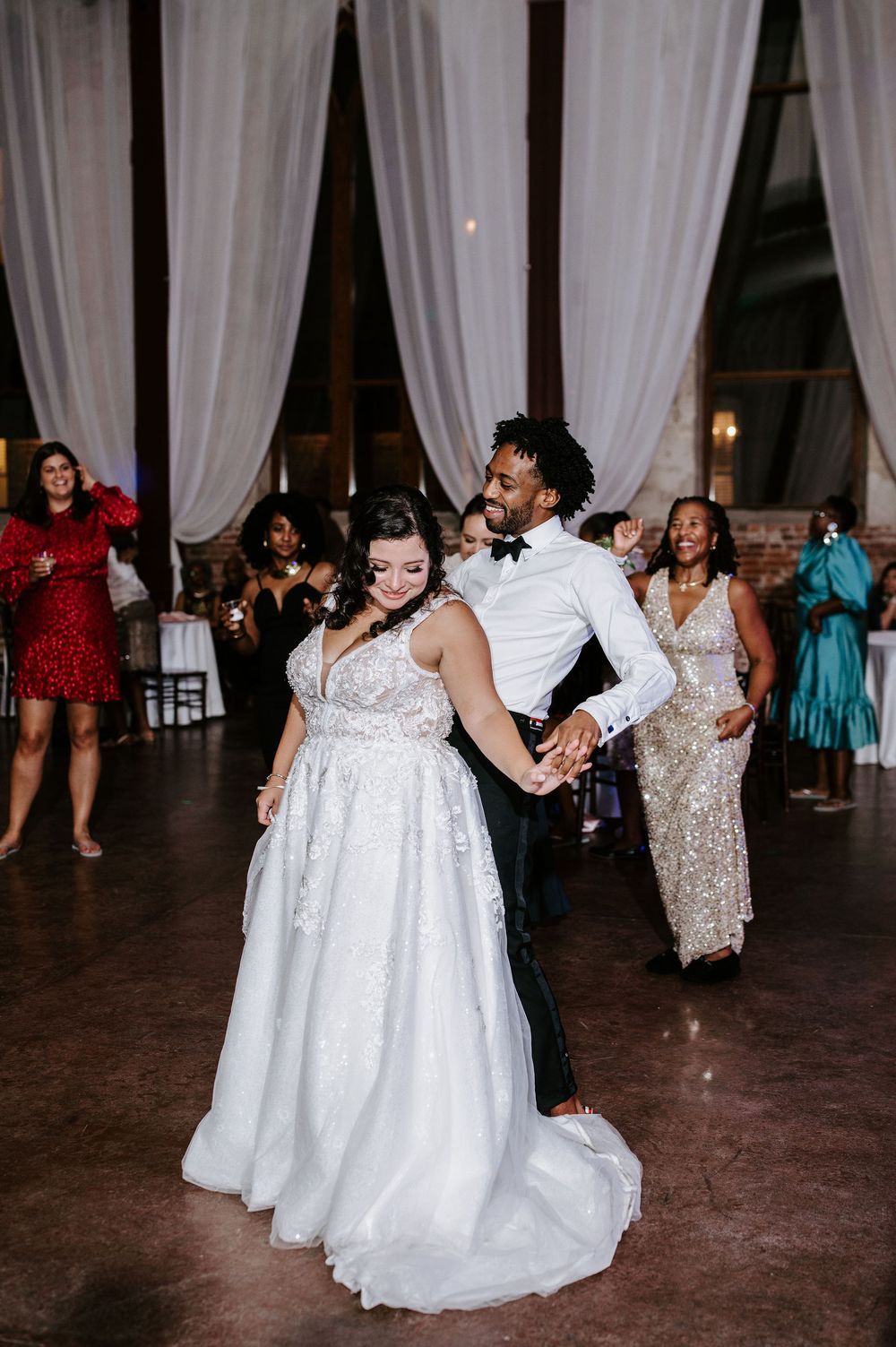 Guests dancing and celebrating at a wedding reception with white curtains and elegant decor in the background.