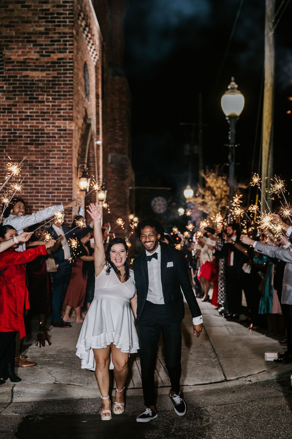 Wedding guests hold sparklers during an evening send-off celebration outside a brick building.