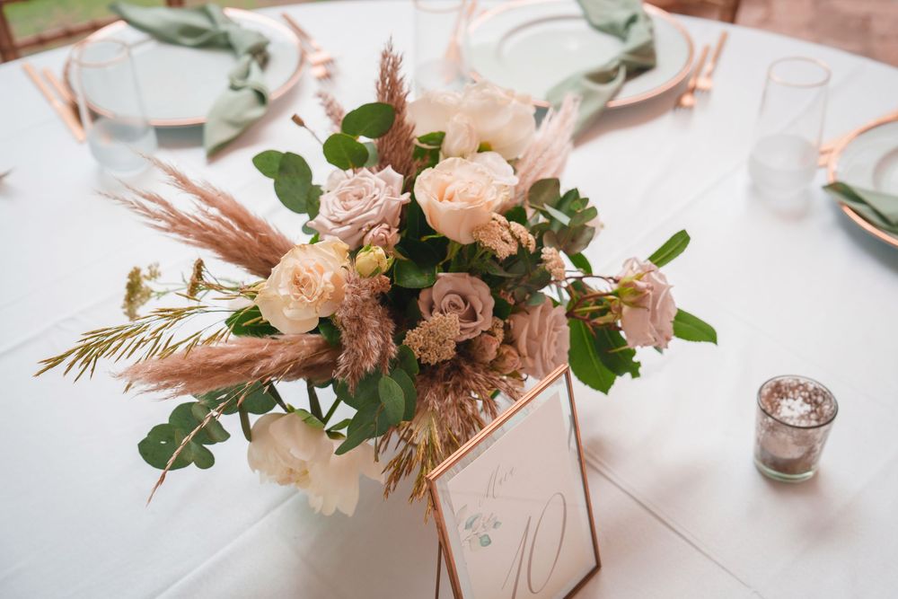 Elegant floral centerpiece with blush roses and dried pampas grass on white tablecloth with place settings.