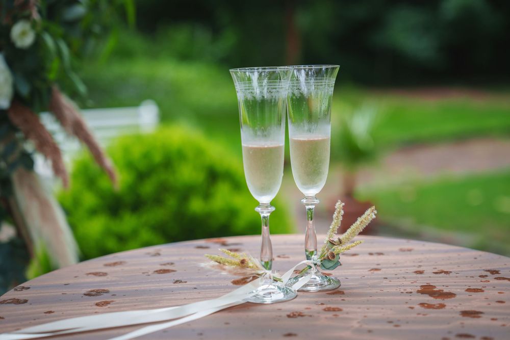 Two champagne flutes filled with bubbly drinks sit on a rustic wooden table in an outdoor garden setting.