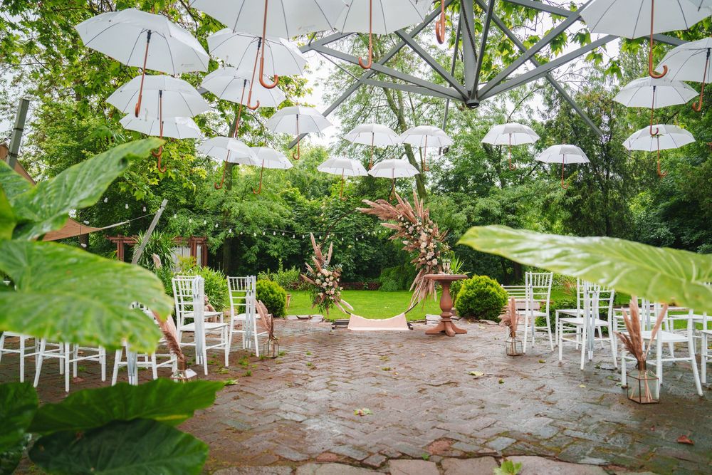 White umbrellas and chairs decorate an outdoor garden ceremony space with stone patio and floral arch.
