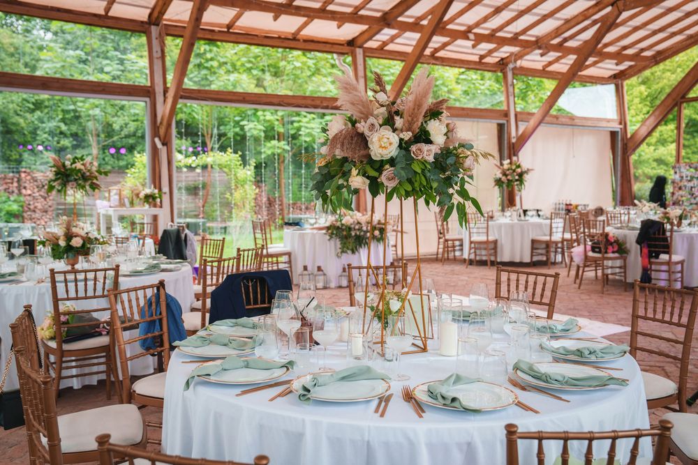 Elegant outdoor reception setup with round tables, chiavari chairs and hanging greenery under a covered pavilion.