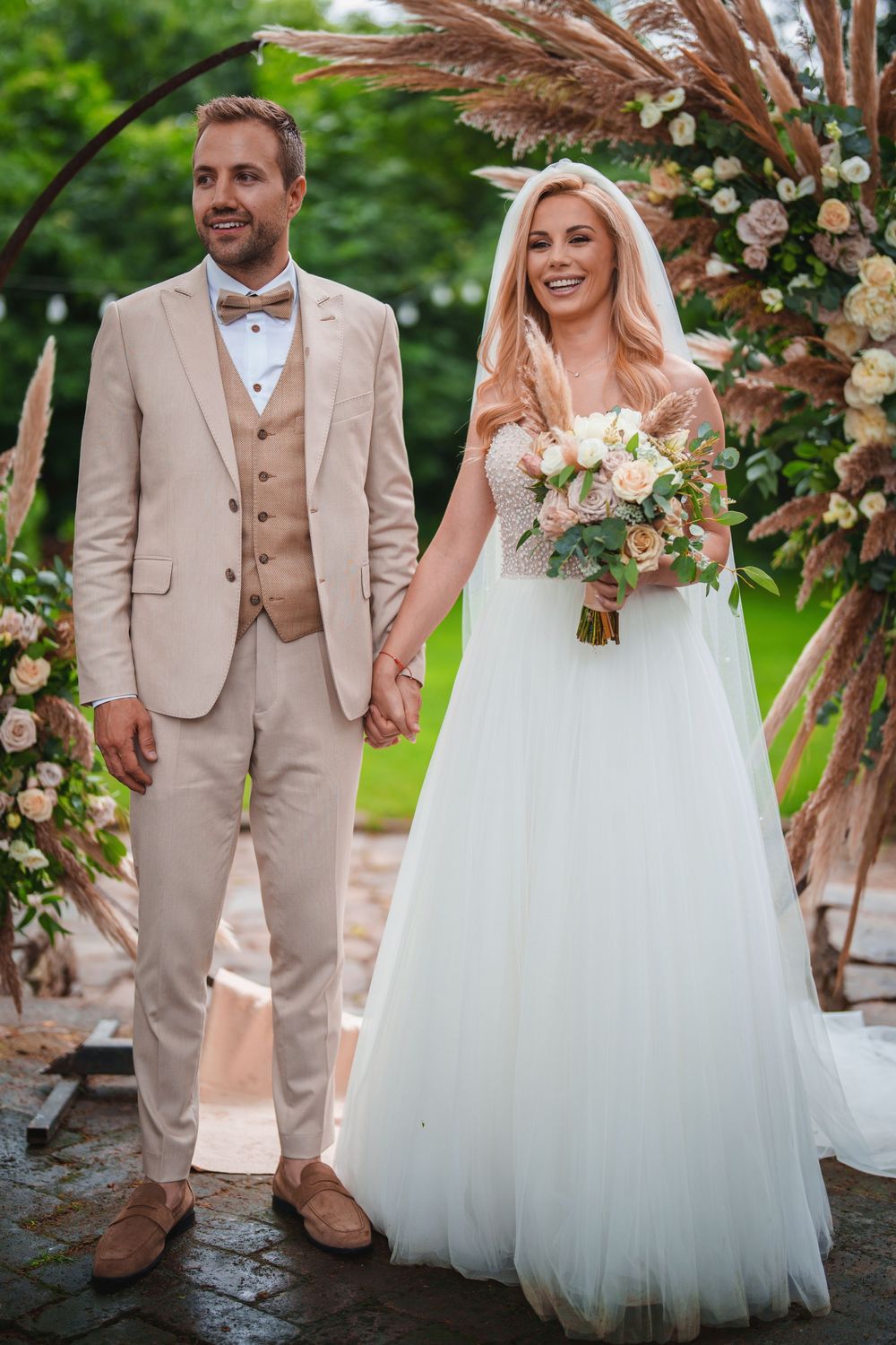 Wedding couple in beige suit and white gown pose outdoors with floral arch and greenery backdrop.