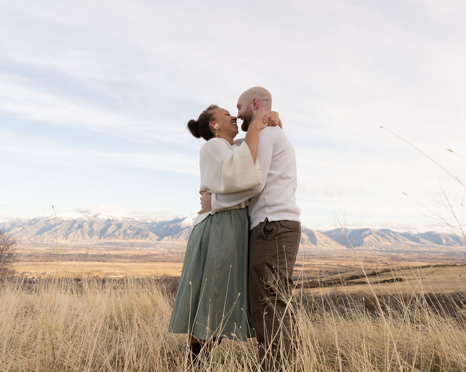 Engagement photo in Cache Valley, Utah, featuring a couple hugging in a scenic outdoor field with the Logan Mountains in the background