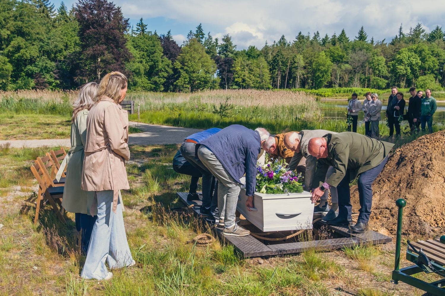 Een kist wordt neergelaten in een graf op een natuurbegraafplaats. 6 mensen staan rond de kist. De uitvaartleiders kijken toe.