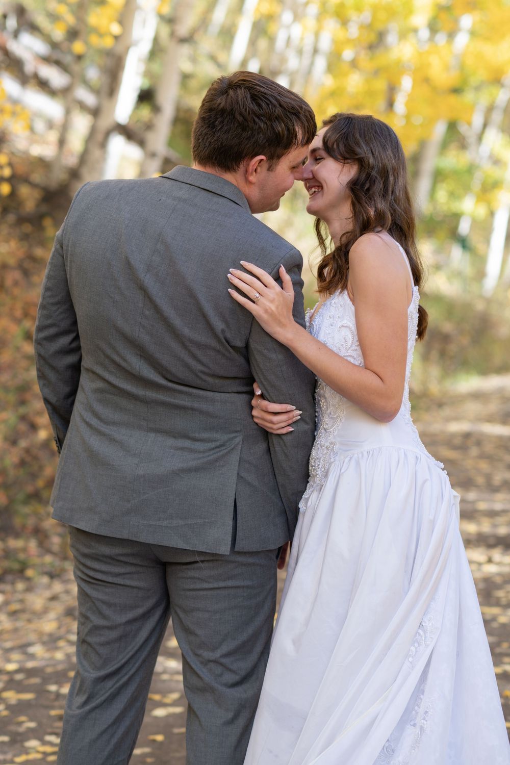 Bride and groom share a romantic moment outdoors in autumn, surrounded by golden trees, as she smiles in a white gown.