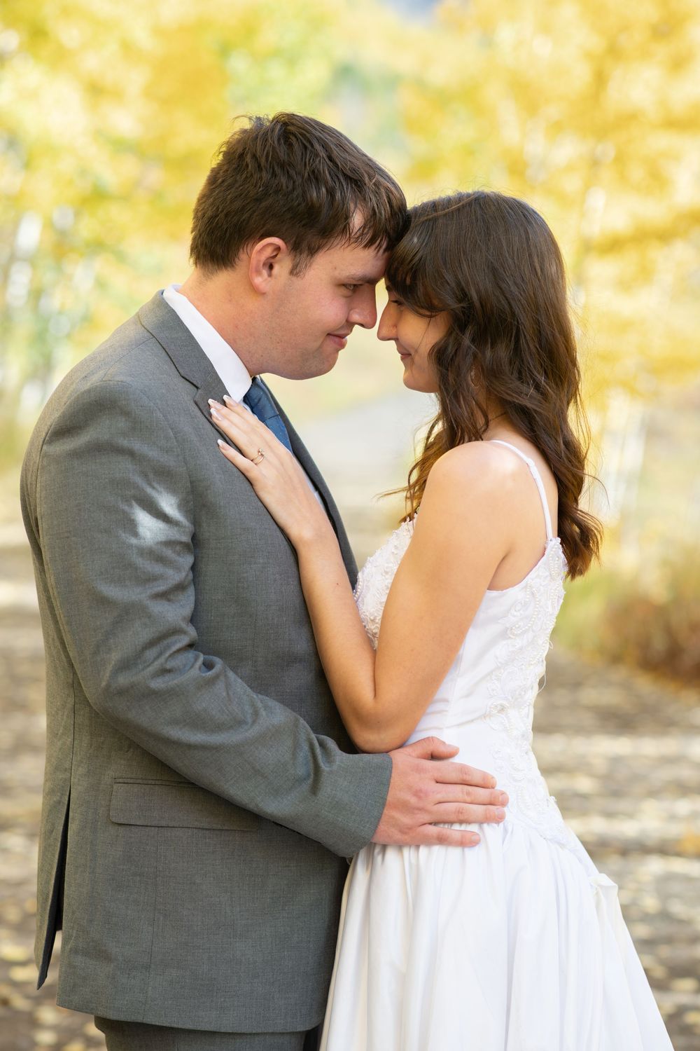 A couple in wedding attire embraces tenderly outdoors, surrounded by golden autumn trees in a romantic fall setting.