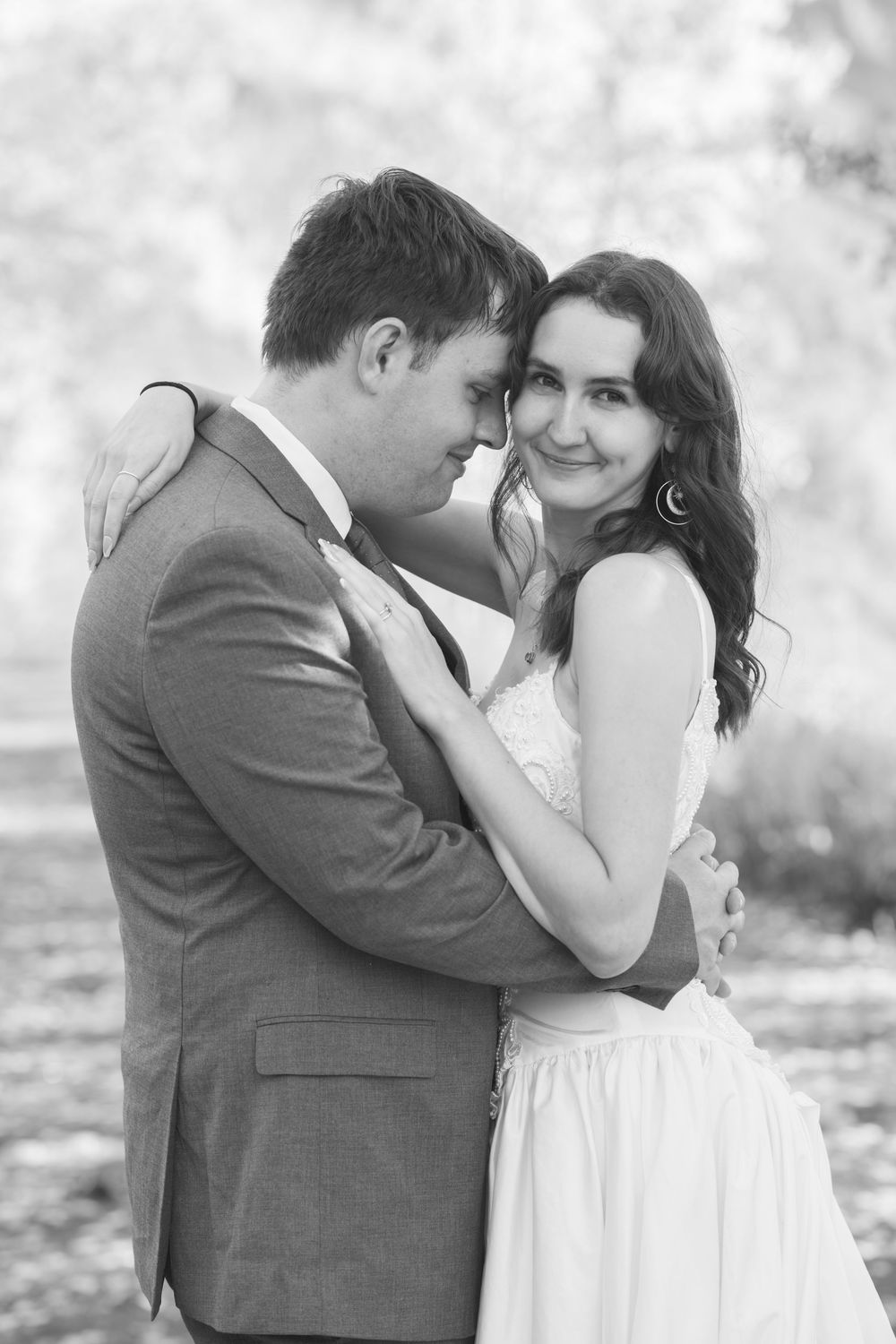 Black and white photo of a bride and groom embracing outdoors, bride in white dress and groom in gray suit.