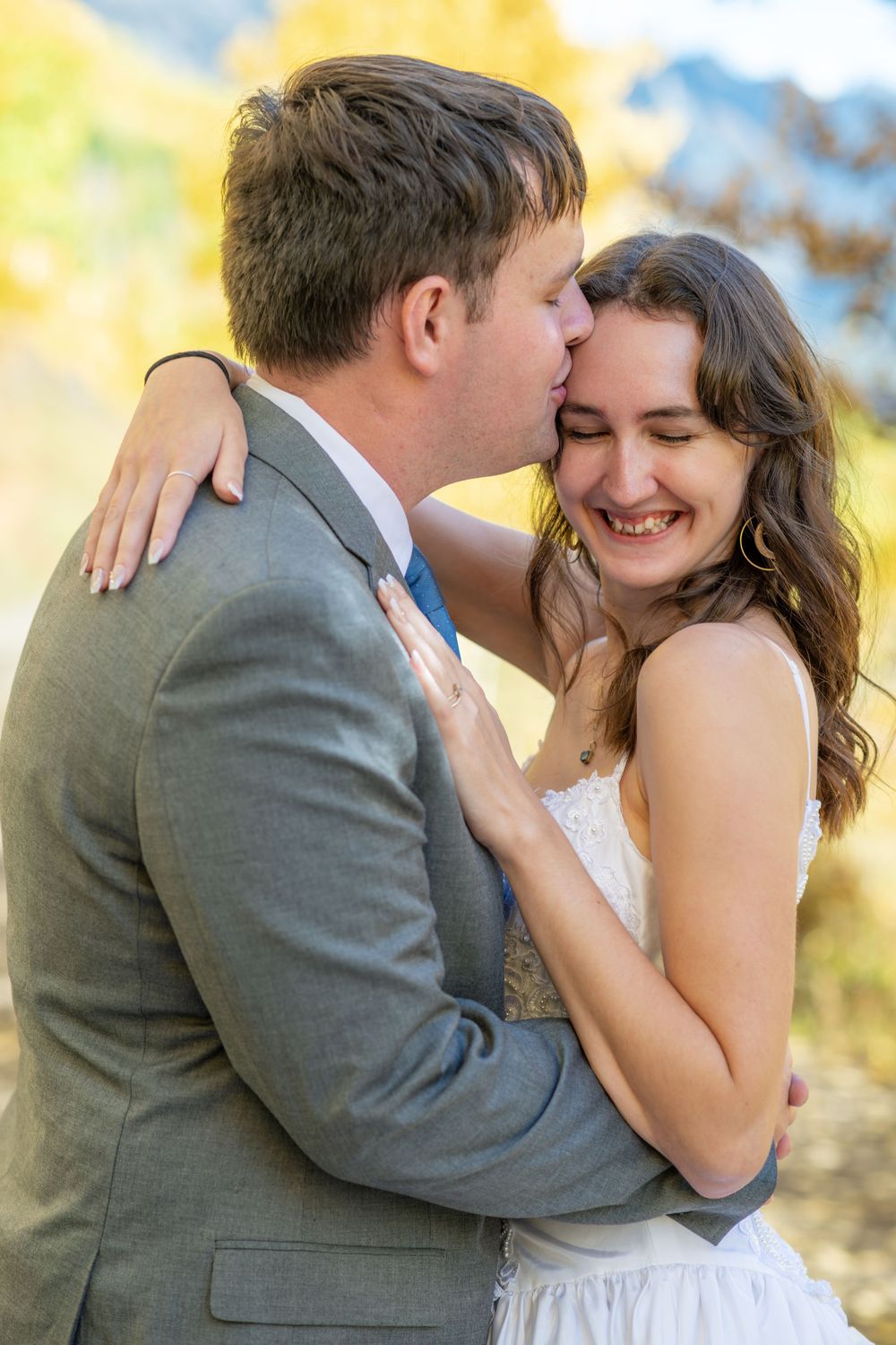 Groom in gray suit kisses bride on head as she smiles joyfully, embracing outdoors with autumn foliage backdrop.