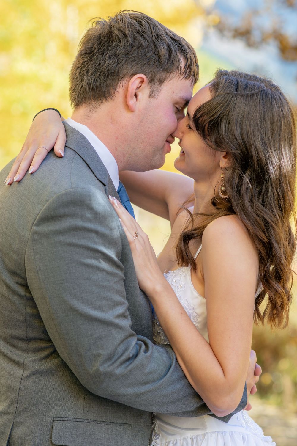 A bride and groom share a romantic moment, nearly kissing outdoors in autumn light, dressed in wedding attire.
