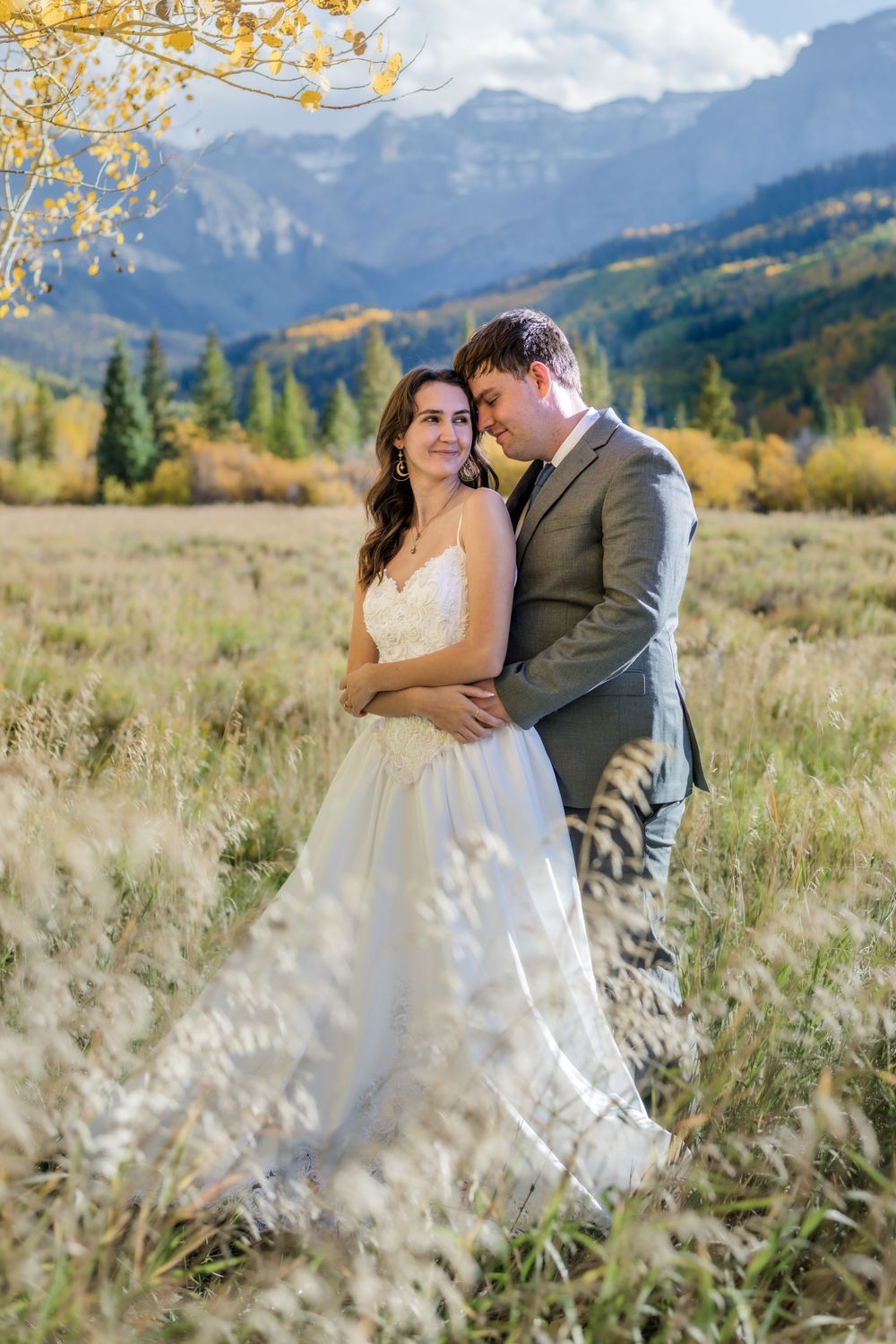 Bride and groom embrace in a golden autumn meadow with majestic mountain peaks and colorful fall foliage in the background.