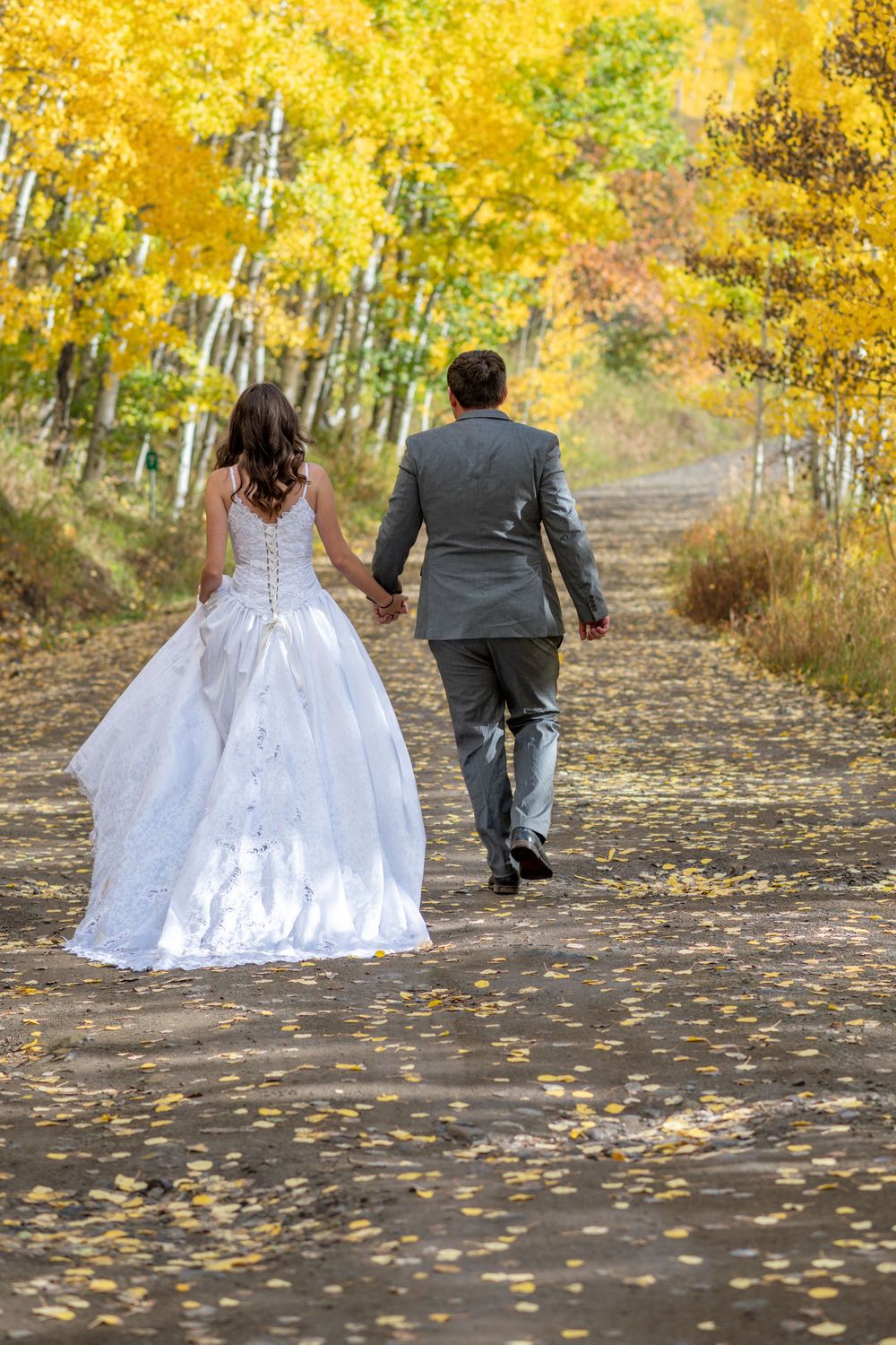 Bride and groom walking hand in hand down a scenic mountain road lined with golden aspen trees in the fall near Ridgway.
