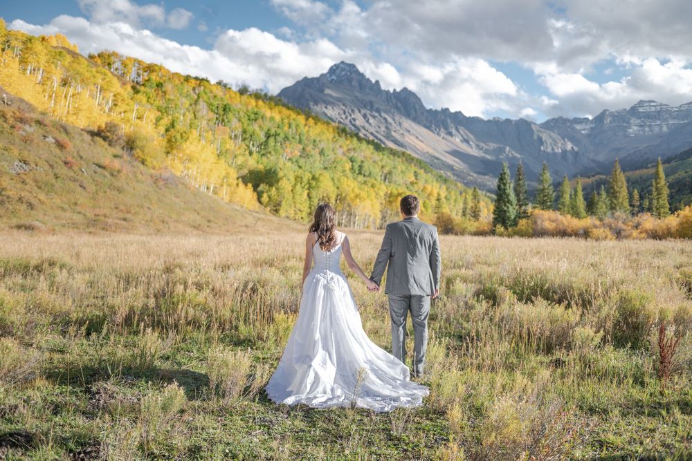Bride and groom holding hands in a mountain meadow with colorful autumn trees and rugged mountain peaks surrounding them.