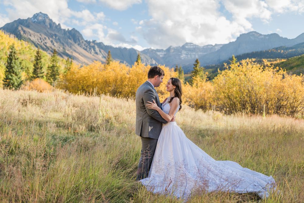 A bride and groom embrace in a mountain meadow with golden autumn foliage and rugged peaks under a blue sky in the San Juan Mountains