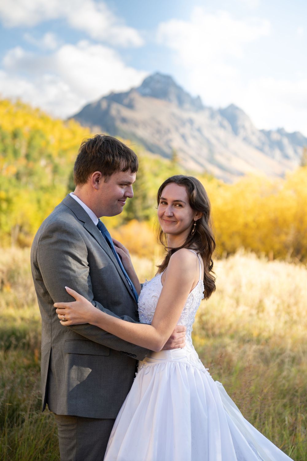 A bride and groom embrace in a mountain meadow with golden autumn foliage and rugged peaks under a blue sky near Ridgway.
