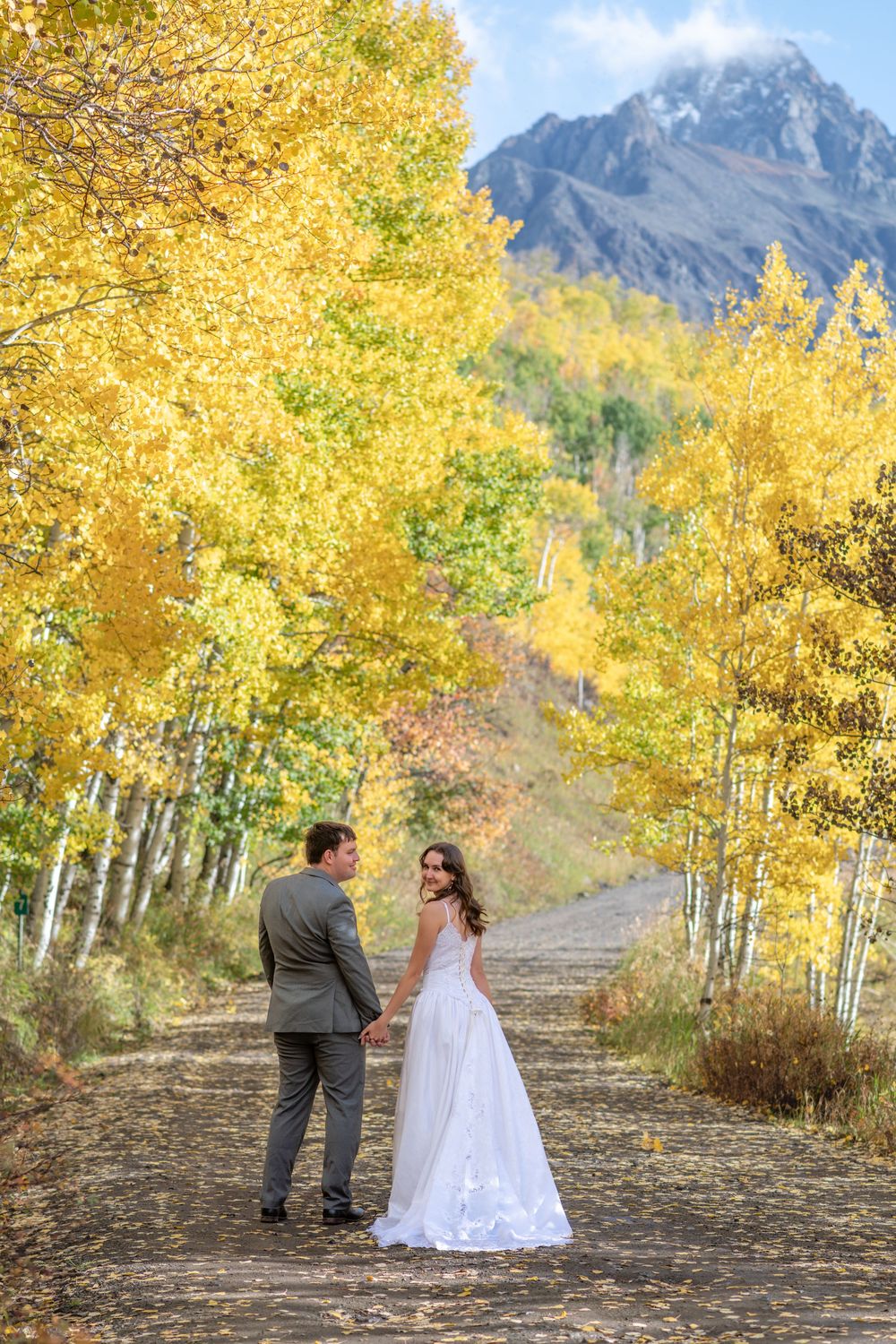 Bride and groom hold hands on a scenic autumn path lined with golden aspen trees and a mountain peak with clouds in the background.