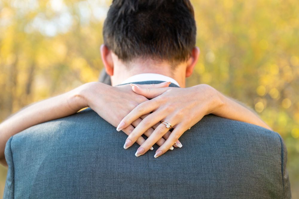 A detail image of a bride's hands around her groom's shoulders showing off her wedding ring with golden aspen trees in the background.