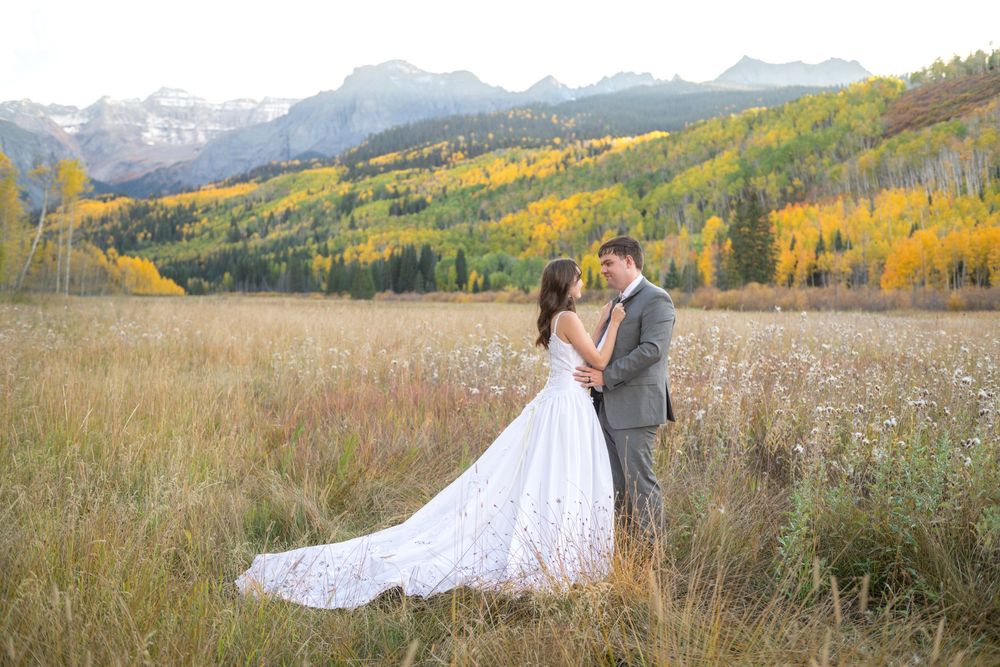 Bride and groom embrace in a golden autumn meadow with colorful aspen trees and mountain peaks in the background.