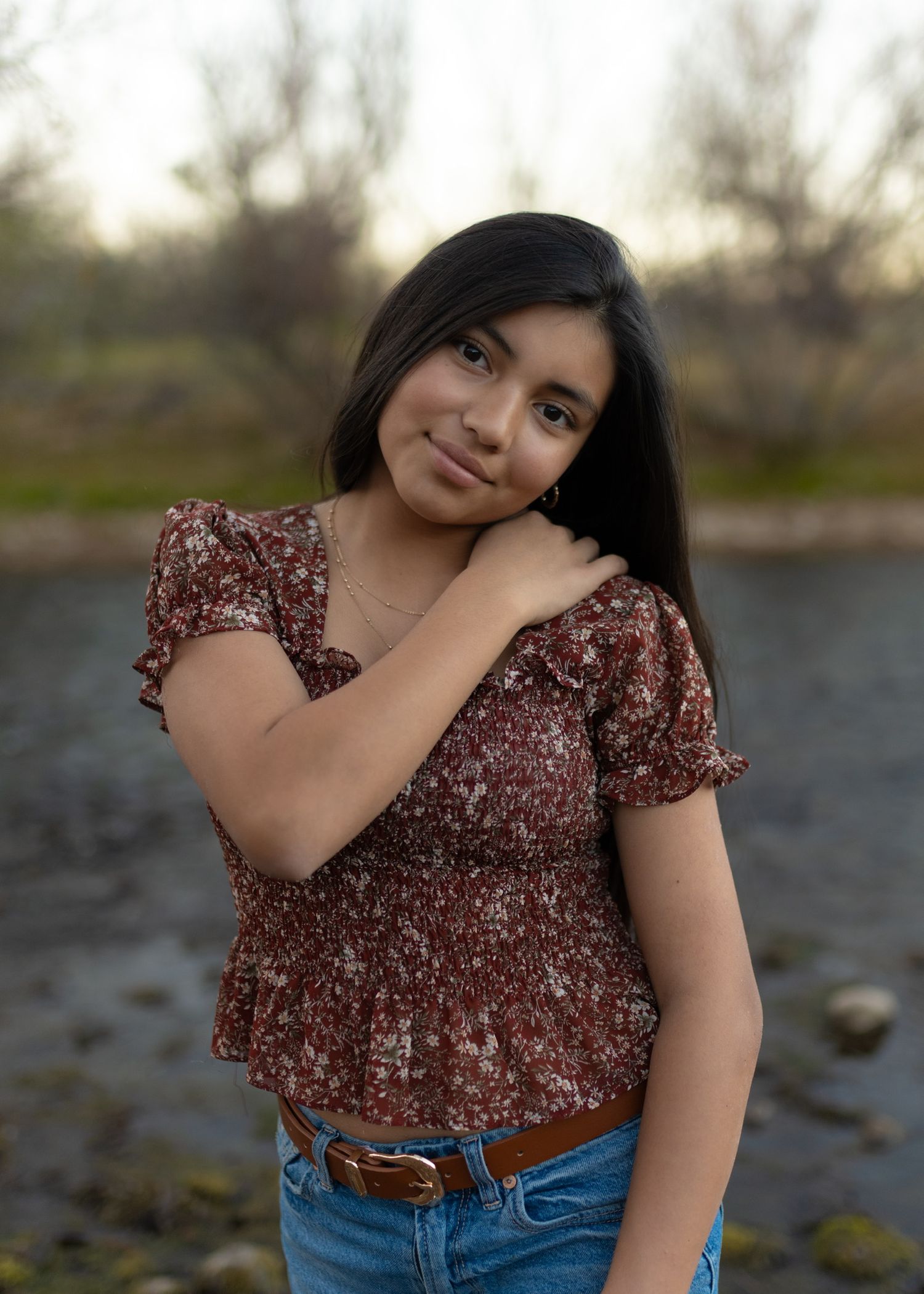 Woman in floral top and jeans poses by water during outdoor photoshoot.