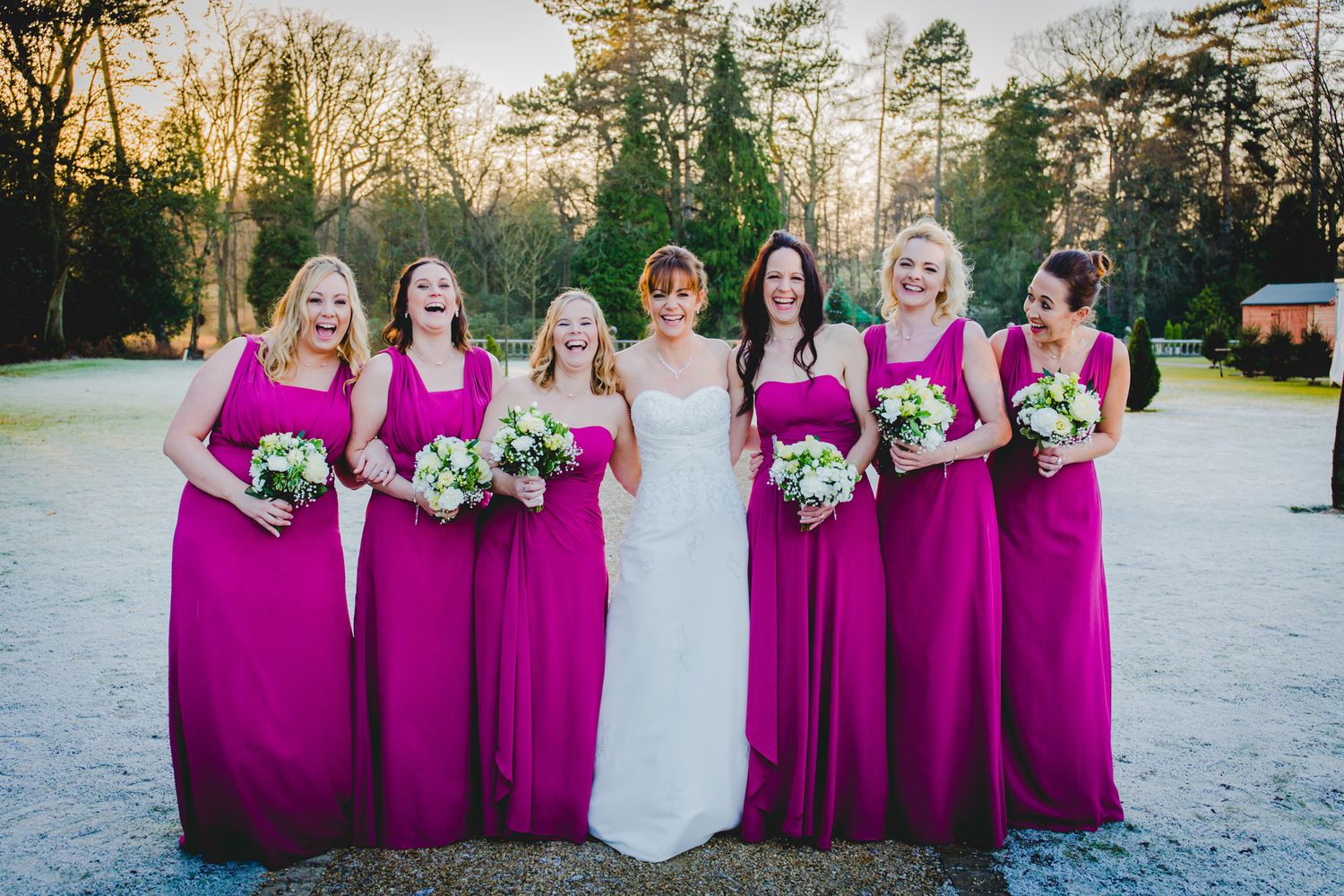 A wedding party poses outdoors in bright magenta bridesmaid dresses and formal attire at sunset on a winter day.