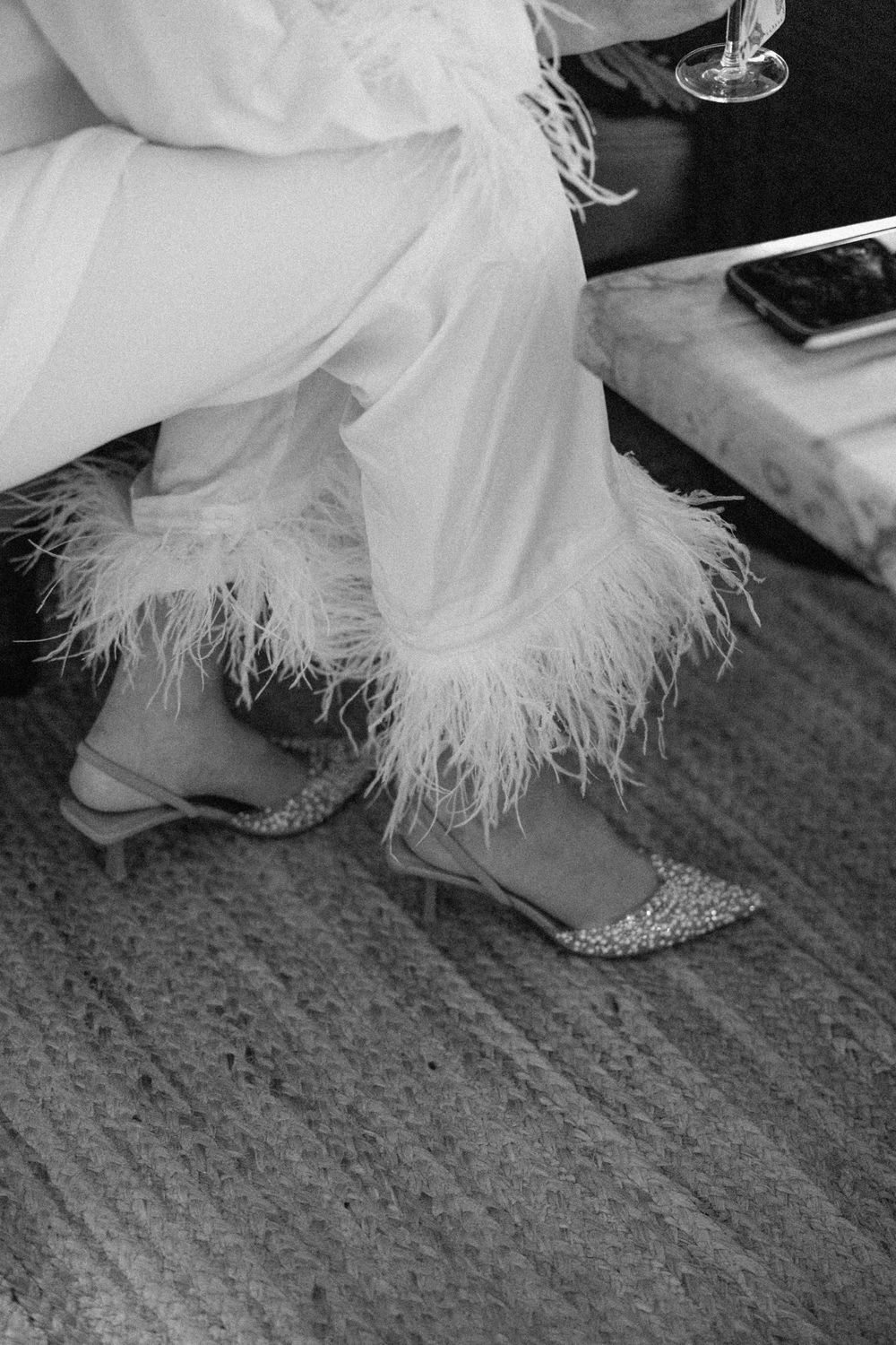 Close-up of feather-trimmed wedding dress hem and shoes on textured carpet.
