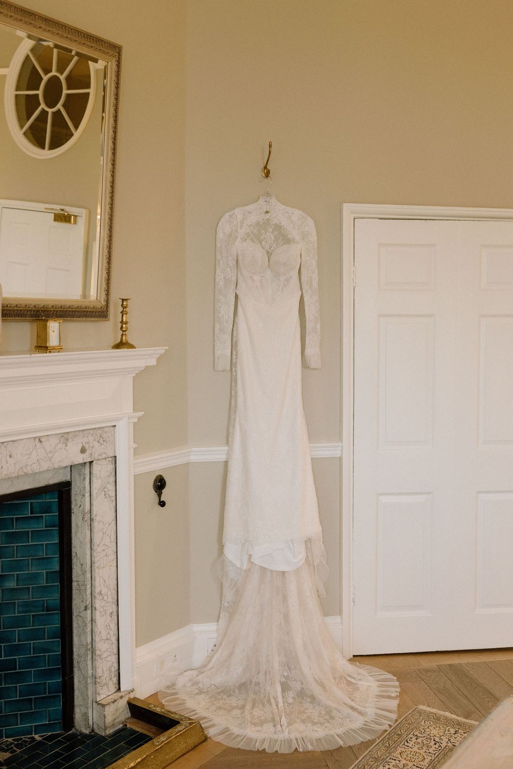 Elegant lace wedding dress with long sleeves hanging on display against cream walls near a marble fireplace.