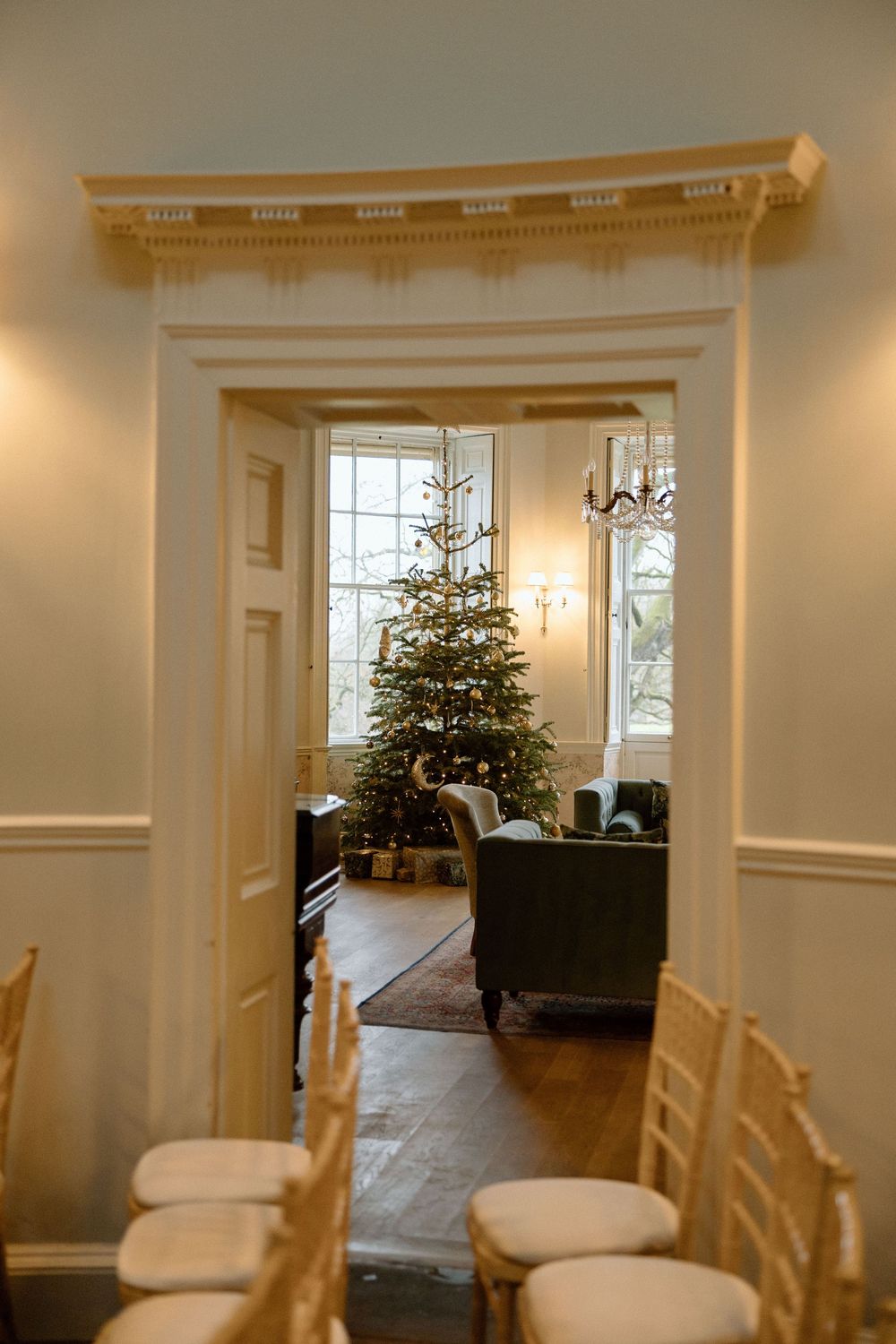 A decorated Christmas tree is visible through an elegant doorway with white trim and wooden chairs lining the hall.
