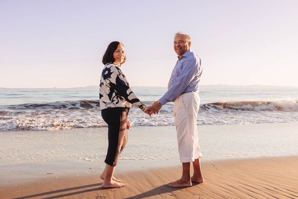 Two people hold hands while standing on a sandy beach at sunset with ocean waves in the background.