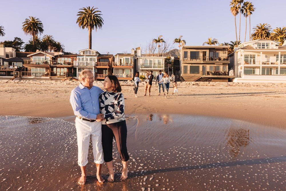 Couple walking along a scenic beach at sunset with palm trees and beachfront houses in the background.